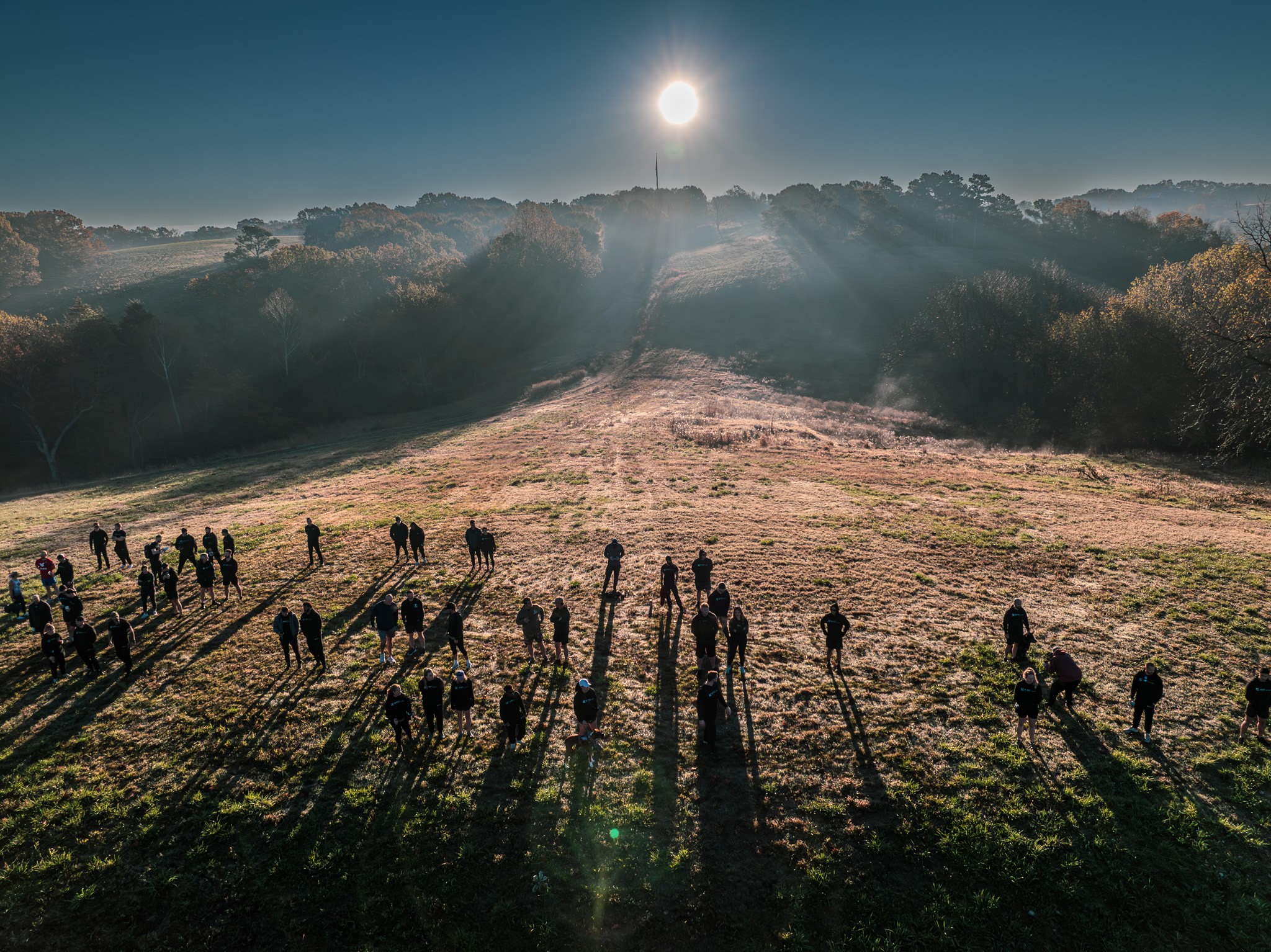 7ecae54091af35aef6458af51e1bdb9ad0a0cddd A group of people stand scattered across a sunlit field at sunrise, casting long shadows on the grass with trees, rolling hills, and a bright sun in the background.
