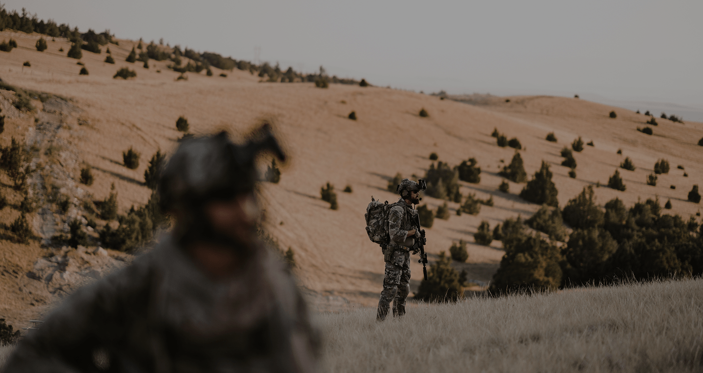 about-us-hero Two soldiers in camouflage gear stand in a grassy, hilly landscape with sparse trees. One is in focus in the distance while the other is blurred in the foreground. Both appear alert and equipped for a mission.